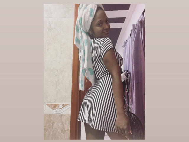 MelanyScoth with dark skin, wearing a striped dress, towel on head, looking back, showing her ass, standing in a bathroom with a purple curtain.