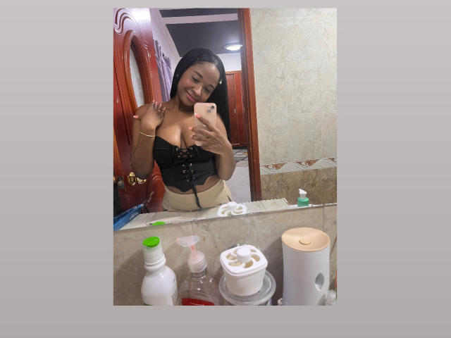 MelanyScoth with dark skin, long black hair, smiling, wearing a black lace-up bustier, holding a phone, mirror selfie, bathroom, countertop with toiletries, beige walls, wooden door.