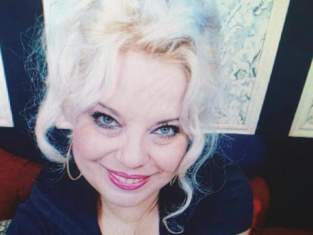 SoniaHotMilf with blonde hair, blue eyes, and pink lipstick, smiling at the camera, wearing gold hoop earrings, in a dark top, with a slightly wrinkled face.