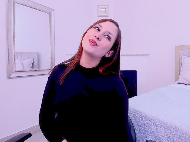 Laia Reyd, con labios rosados y voluminosos, un suéter negro y cabello rojo, inclinada ligeramente hacia atrás, sentada frente a una cama con ropa de cama blanca..
