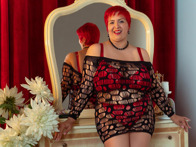LadyLibely with bright red hair, smiling, wearing a black mesh dress revealing red bra and panties, sitting in front of a mirror, red curtains, white flowers, and a wooden dresser.