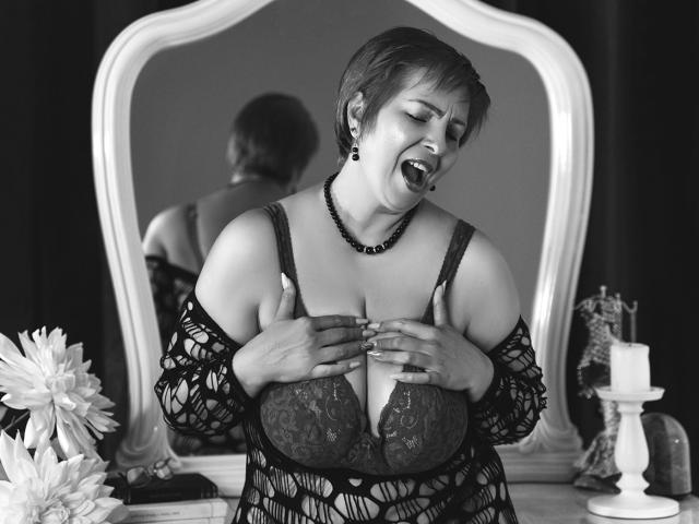 Black-and-white photo of LadyLibely with short hair, wearing lace bra and net dress, hands on her tits, mouth open, necklace, mirror, candle, and flower in background.