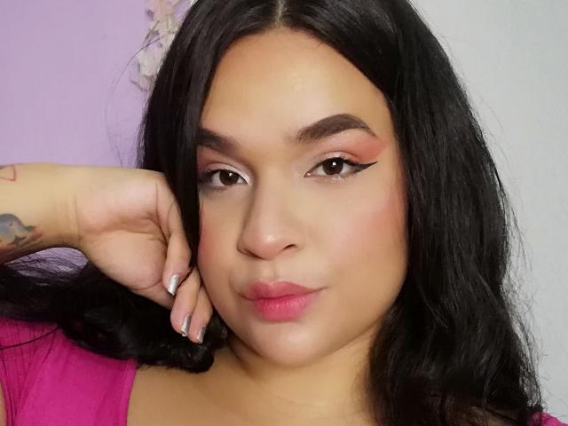 AntonelaPark tem cabelo preto, delineador rosa e batom rosa, com uma tatuagem na mão esquerda, veste uma blusa rosa e tem um leve franzido nos lábios, com um bumbum cheio e arredondado..