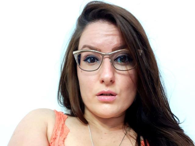 BadAngel with brown hair, glasses, and pink lips, wearing an orange lace top, has a slightly confused or surprised expression. Her tits are small and her ass is unseen.