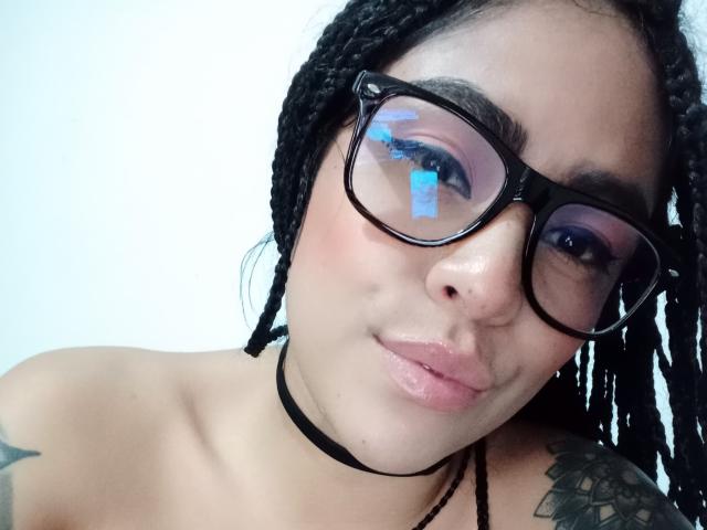 StellaBeckker with black braids, purple eyeshadow, thick black glasses, shiny pink lips, black choker, tattoo on right shoulder, and visible cleavage, smiling slightly.