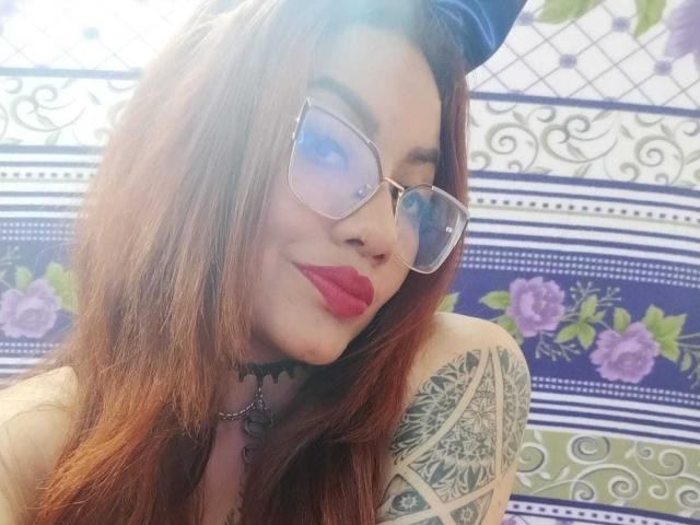 Alexandra Petit, com cabelo ruivo, óculos, batom vermelho, colar preto, tatuagem no ombro direito, piscando, tiara azul, exibindo os seios, sorrindo, parecendo fofa, sexy e confiante..