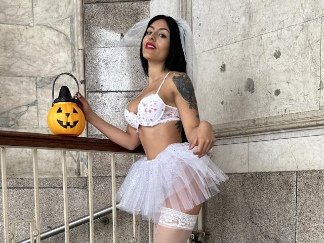DellaSmith vestida con lencería blanca, con un sujetador con estampado floral, una falda de encaje, una liga blanca y un velo, sosteniendo un farol de calabaza, de pie en una escalera de mármol, mostrando un tatuaje en el brazo izquierdo..