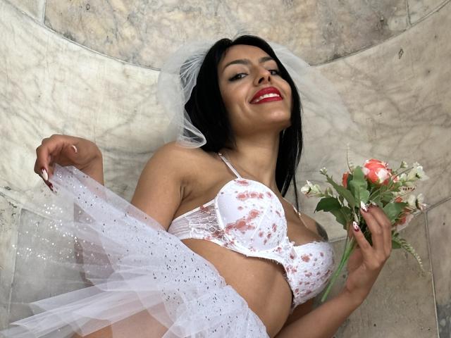 DellaSmith, avec ses cheveux noirs, son soutien-gorge blanc à motifs floraux et son voile, tenant des roses roses, soulevant sa jupe blanche scintillante, souriant avec du rouge à lèvres, sur un fond de marbre, montrant ses seins et tenant des fleurs..
