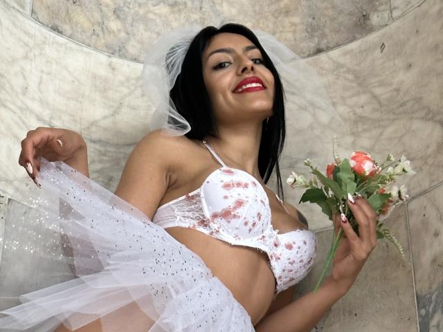 Della Smith, con i capelli neri, indossa un reggiseno bianco con fiori, tiene un bouquet, solleva una gonna bianca scintillante, sorride con il rossetto rosso, e ha un velo sulla testa..