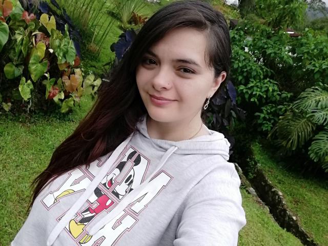Katherin, een Latina met lang bruin haar, lichte huid, kleine borsten, draagt een grijze hoodie met Mickey Mouse erop, ze glimlacht, staat op groen gras, haar oorbellen zijn zichtbaar, en ze kijkt naar de camera..