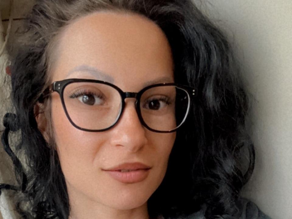 EmilyKross69, met zwart, krullend haar, bruine huid en zwarte bril, toont een subtiele glimlach. Ze heeft een schattig gezicht en haar borsten zijn niet zichtbaar..