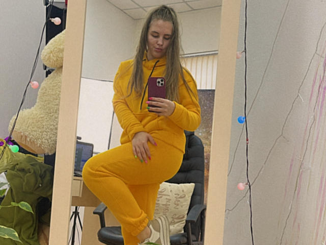 Dooris with long blonde hair, yellow hoodie, and matching yellow pants, standing in front of mirror, right leg crossed, holding red phone, in office, wearing white sneakers.