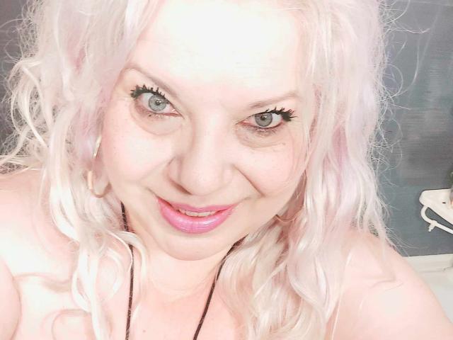 SoniaHotMilf has pale pink curly hair, green eyes, and pink lipstick, smiling at the camera with long black eyelashes, wearing a black necklace, showing her bare shoulders.