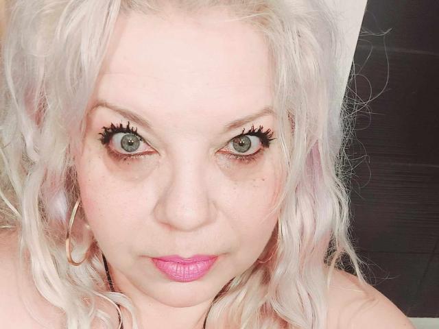SoniaHotMilf with platinum blonde hair, green eyes, and pink lipstick, wearing hoop earrings, staring intensely with long, dark eyelashes, and a slightly parted pink-lipped mouth.