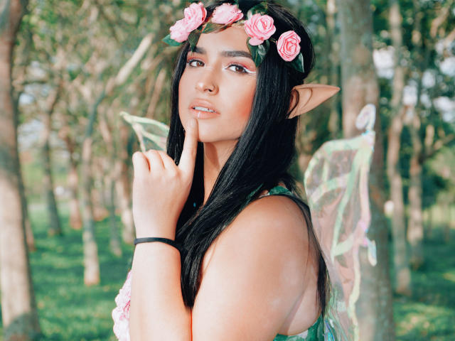 MollyBron with long black hair, pink flower headband, elf ears, and fairy wings, touching her finger to her lip, looking seductive, wearing a green dress.