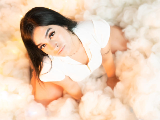 MollyBron, con cabello oscuro, piercing en la nariz y camisa blanca, arrodillada en una nube de humo blanco, mirando hacia arriba con delineador de ojos y una sonrisa sutil, mostrando sus senos y un tatuaje..