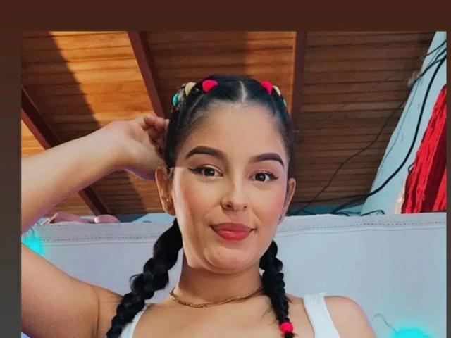 TalianaSquirt with braided black hair, pink and red hair ties, gold necklace, white top, and pouty pink lips, holding her hair up, smiling. Wooden ceiling in background.