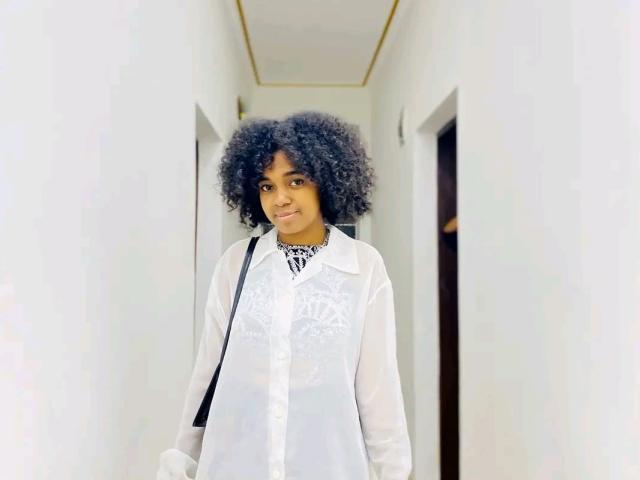 Ariana Delice, con cabello negro rizado, vestida con una camisa blanca de encaje, sonriendo, de pie en un pasillo blanco, con una correa negra sobre el hombro, luciendo linda y elegante..