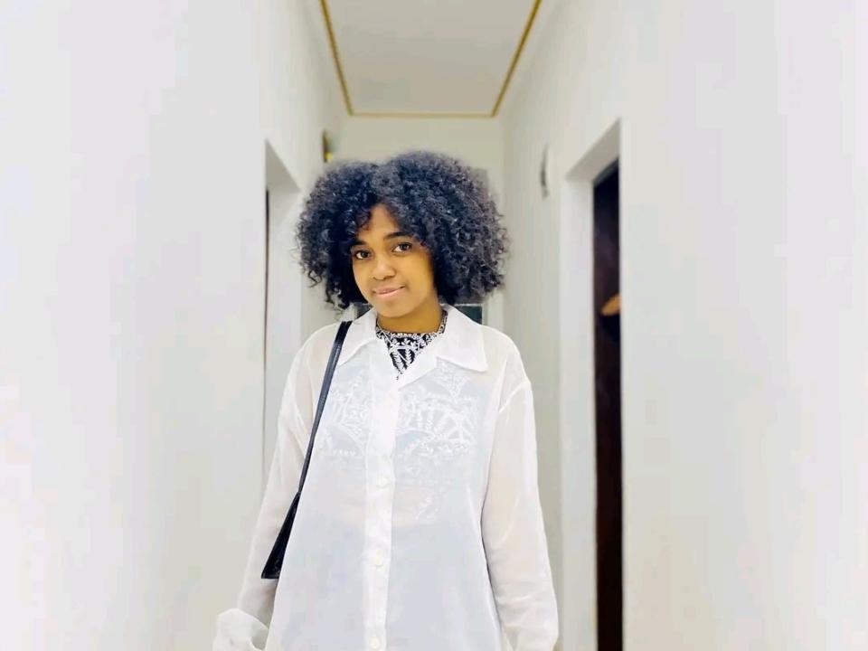 ArianaDelice with curly black hair, wearing a white lace shirt, smiling, standing in a white hallway, black strap over shoulder, looking cute and stylish.