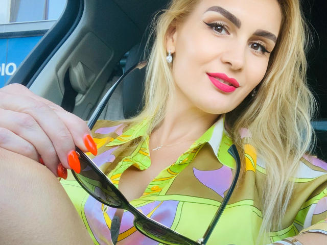 Een aantrekkelijke vrouw met blond haar, roze lippenstift en oranje nagels, gekleed in een kleurrijk shirt, die een zonnebril vasthoudt, zittend in een auto, en haar borsten laat zien..