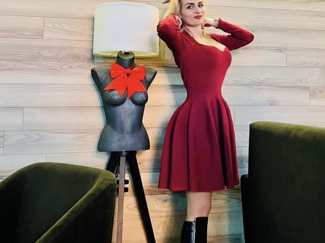 Una mujer atractiva con un vestido rojo, de pie con las manos detrás de la cabeza, al lado de un maniquí gris con un lazo rojo, en una habitación con sillas verdes y una pared de madera..