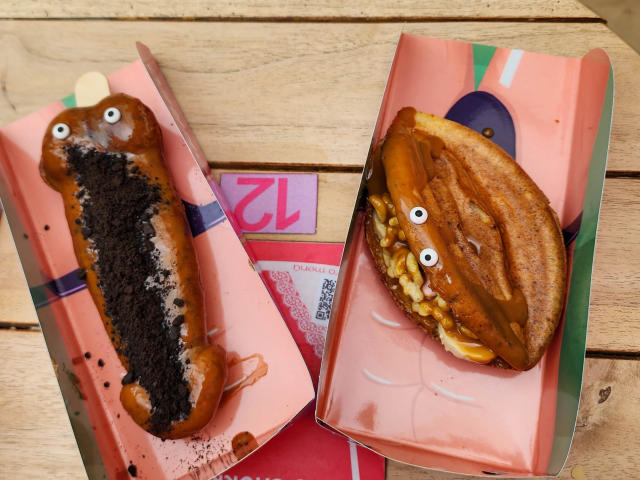 Twee grappige hotdogs met ogen, waarvan er één bedekt is met chocolade-Oreo kruimels en de andere met karamel en mais, verpakt in roze papieren wikkel, op een houten tafel. JainaRogers heeft deze gemaakt..