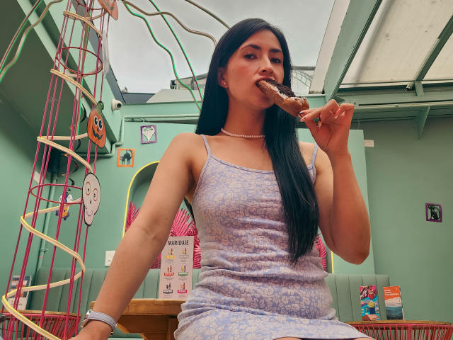 JainaRogers with long black hair, eating a donut, wearing a light purple, floral dress, and a pearl necklace, sitting in a colorful room. Her tits are small.