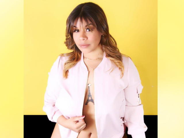 Valeri Harris, avec ses cheveux bruns ondulés, porte une chemise rose ouverte, révélant un tatouage sur sa poitrine et un aperçu de ses parties intimes. Fond jaune..