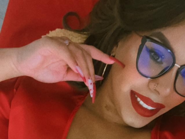 Valeri Harris, avec du rouge à lèvres, un anneau de nez et des lunettes, tenant une carte rose avec des points rouges, portant une chemise rouge, souriante, avec des ongles longs, des cheveux noirs ondulés, en gros plan, détendue..