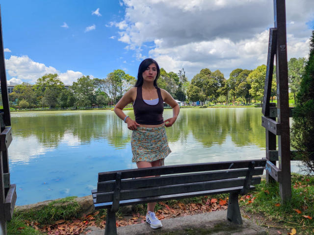 JainaRogers, with small tits, stands by a lake in a black tank top and colorful skirt, hands on hips, in front of a wooden bench. She's wearing white sneakers.