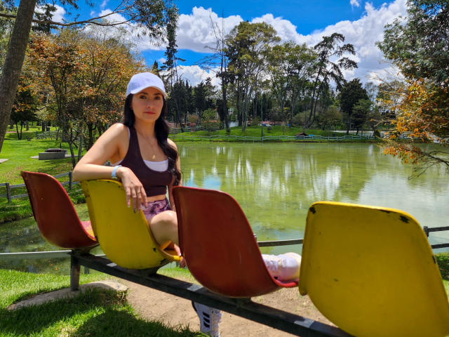 JainaRogers with tits covered by a dark tank top, wearing a white cap, sitting on yellow and red plastic chairs by a green pond, sunny day, grassy park.