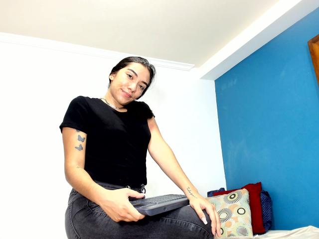 SharonKlart with black shirt and black jeans, pulling down pants, smiling, showing tattoos on arms, dark hair, blue wall, white ceiling, and red and patterned pillows in background.