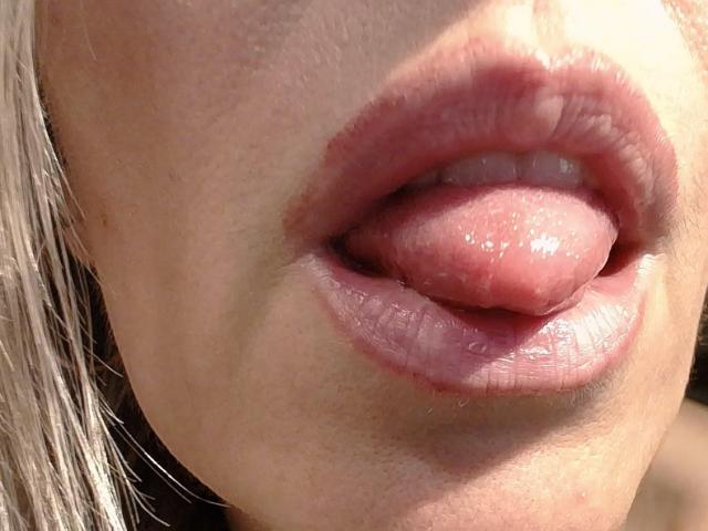 La lingua di VickySexyFr sporge dalle sue lucide labbra rosa, mostrando un po' dei suoi denti superiori e un po' di saliva sulla lingua. La sua pelle è chiara..