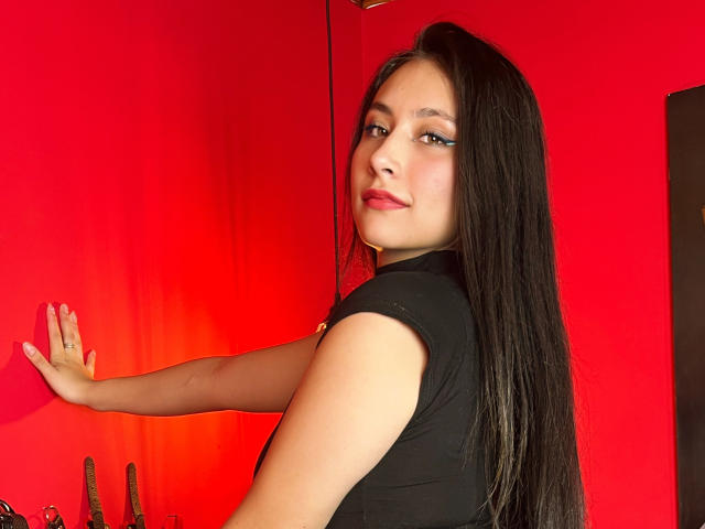NiobeSmith, avec ses longs cheveux noirs, son rouge à lèvres rouge et son haut noir, se tient devant un mur rouge, regardant par-dessus son épaule, une main sur le mur, un léger sourire, sexy, attirante..