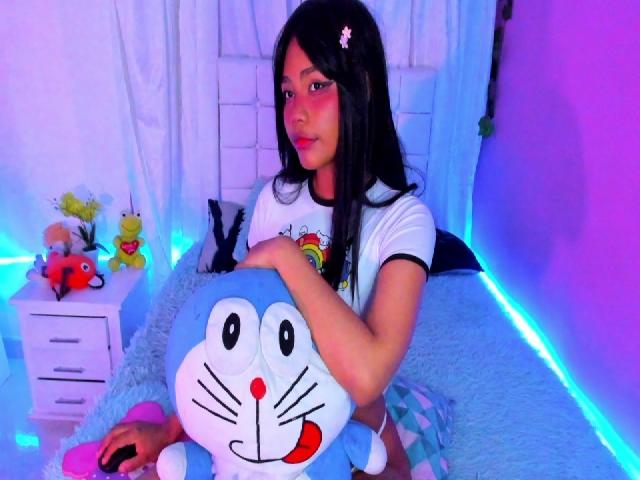 AnniieeBell with long black hair, wearing a white shirt, hugging a blue Doraemon plushie, sitting on a blue bed, surrounded by stuffed animals, in a purple-lit room.