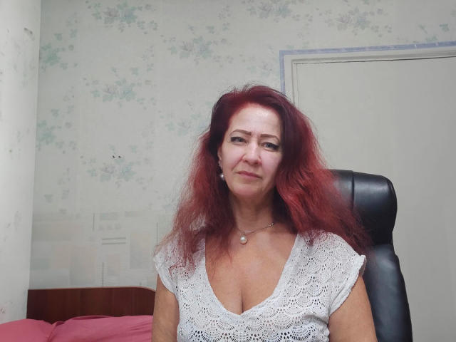 MilayaDFlow mit roten Haaren, einem weißen Spitzenoberteil, das ihre Brüste zeigt, sitzend auf einem schwarzen Stuhl, mit einer Perlenkette um den Hals, leicht lächelnd, in einem einfachen Raum..