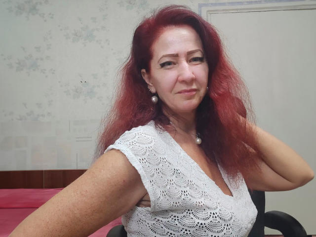 MilayaDFlow has red hair, white lace top, pearl earrings, and a slight smile. She's sitting in a chair with a pink pillow behind her.