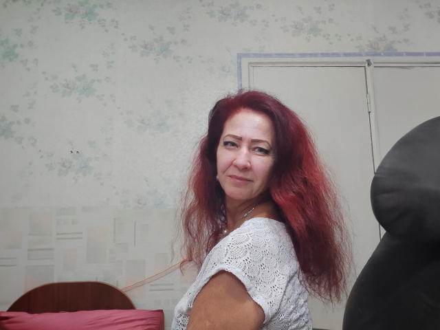 MilayaDFlow, de meia-idade, com cabelo ruivo e desalinhado, vestindo uma blusa branca de renda, com um leve sorriso, olhando por cima do ombro, em pé ao lado de uma cadeira preta e um travesseiro rosa..