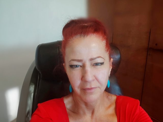 MilayaDFlow a les cheveux roux, des boucles d'oreilles bleues et une chemise rouge, assise sur une chaise noire avec un visage légèrement flou, qui semble fatiguée et porte une expression discrète de contrariété..