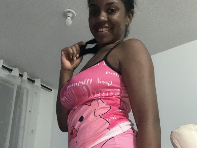 TiffanySammy with dark skin, wearing a pink Hello Kitty tank top and shorts, holding a black hair tie, smiling, showing her tits and ass, in a dimly lit room.