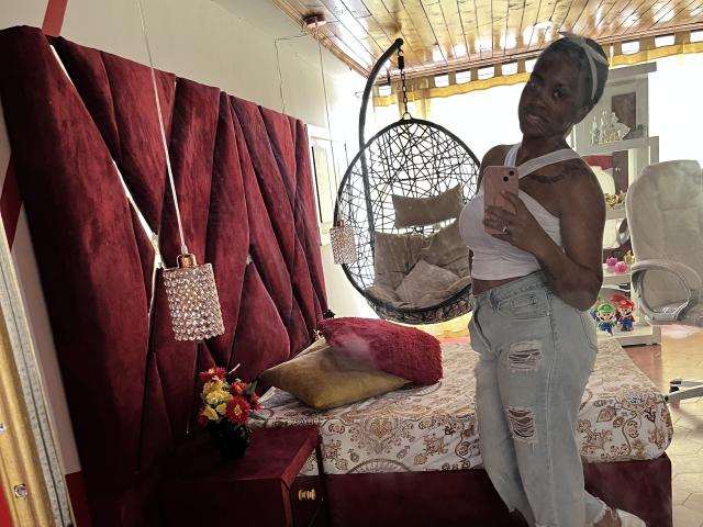 TiffanySammy with dark skin, short gray hair, small tits, white tank top, ripped jeans, standing in bedroom, holding phone, red velvet headboard, hanging chair, floral pillow.