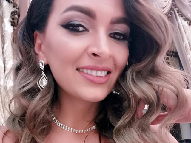 PassionX has wavy blonde hair, dark eyeliner, pink lipstick, and is smiling. She's wearing diamond earrings and a necklace. Her tits are not visible in this shot.