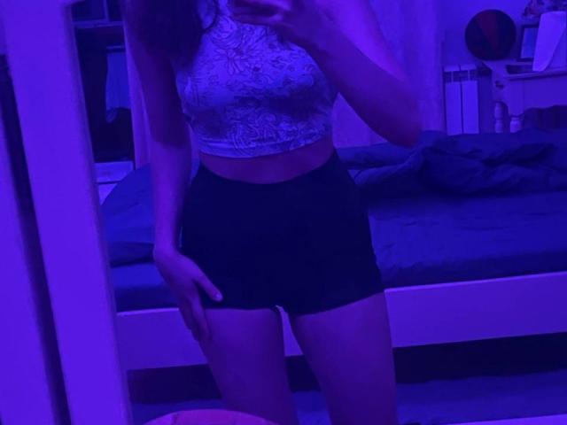 Lukreciaa, dans un autoportrait éclairé en violet, portant un crop top blanc en dentelle et un short noir moulant, montrant sa poitrine et ses fesses, tenant son téléphone dans sa main droite..