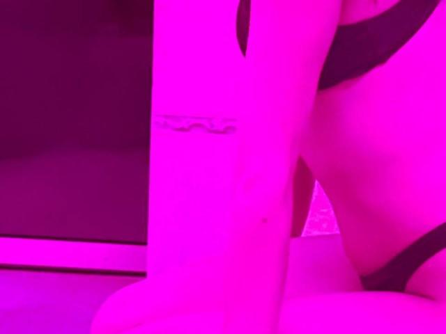 O braço de Lukreciaa com uma pequena tatuagem, usando roupa íntima preta, ajoelhada com o bumbum e a vulva à mostra, tudo sob iluminação rosa neon..