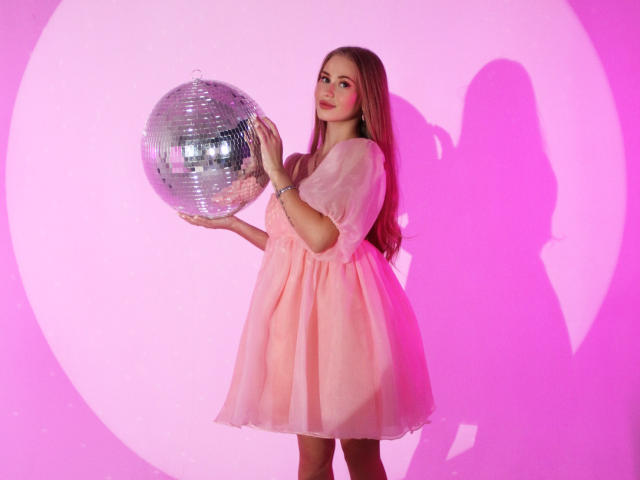 Violetta Steward, con cabello rosa largo, vestida con un vestido de tul rosa, sosteniendo una brillante bola de discoteca plateada, sombra rosa en la pared, de pie, linda, sonriendo, iluminación rosa suave..