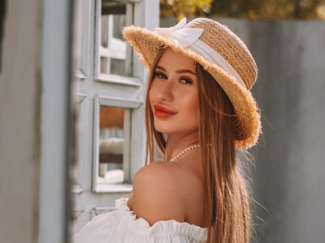 Violetta Steward mit langen, braunen Haaren, die einen Strohhut mit einer weißen Schleife, ein weißes Oberteil mit offener Schulter und eine Perlenkette trägt, lächelt in die Kamera und zeigt ihre freie Schulter und ihren Hals..