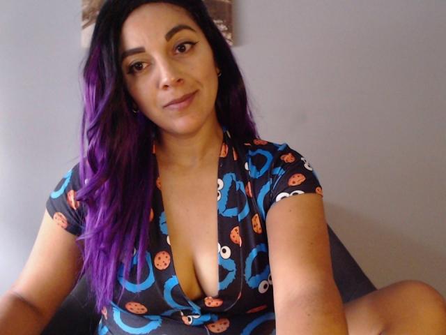 PrettyEllie with purple hair, wearing a blue and orange patterned dress, showing a deep cleavage, sitting, smiling, and taking a selfie, with her legs slightly apart.