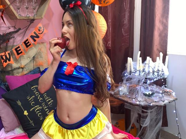 Jessie Warren, avec ses longs cheveux bruns, mange une pomme rouge, vêtue d'une tenue brillante bleue et jaune inspirée de Disney, qui révèle son ventre plat et ses petites seins..