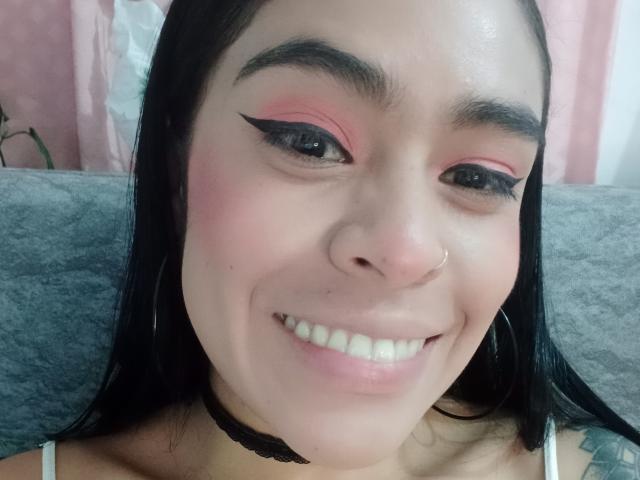 StellaBeckker has pink eyeshadow, black eyeliner, a nose ring, and a black choker. She's smiling with white teeth, black hair, and a tattoo on her right shoulder.