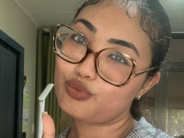 LovelyCrissy, com cabelos castanhos cacheados, usando óculos e com lábios carnudos, segurando um secador de cabelo branco, exibindo seu pout (lábios protuberantes) que lembra um bumbum, e com olhos azuis, em um quarto com iluminação suave..